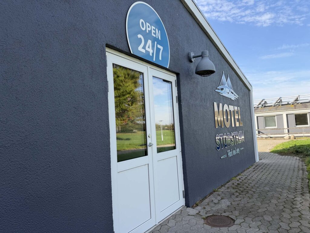 Hvid dobbeltdør med Open 24/7 skilt på mørkeblå facade og motellets logo på muren